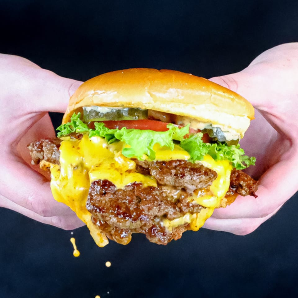 Hands holding a Smashers double cheeseburger with melted cheese, lettuce, tomato, and pickles.