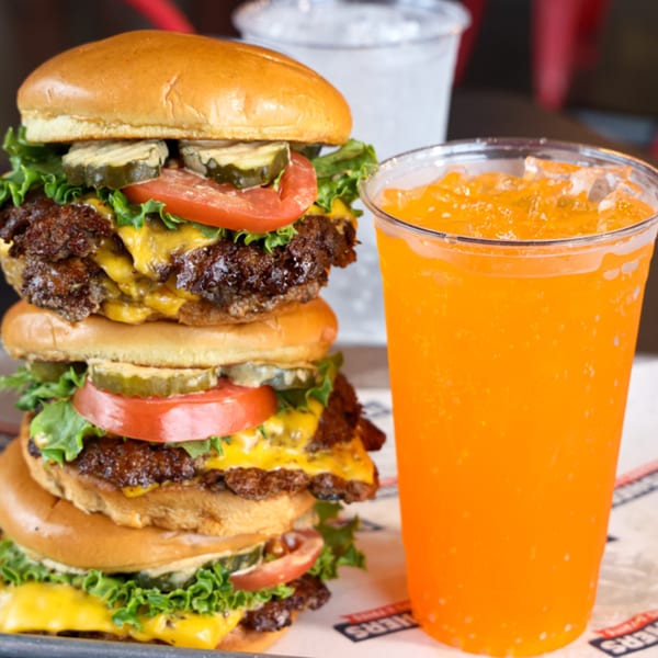 Three stacked Smashers burgers with cheese, lettuce, tomato, and pickles, next to an orange soda.