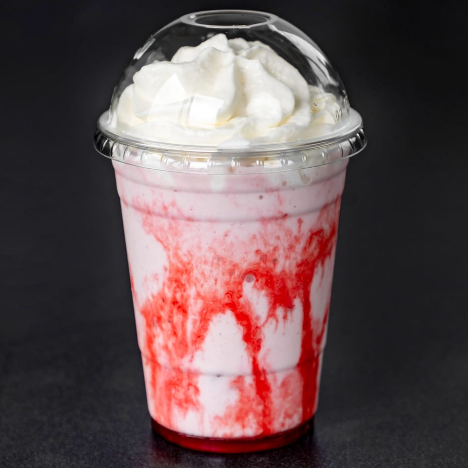 Strawberry milkshake with whipped cream and red syrup swirls in a clear plastic cup.